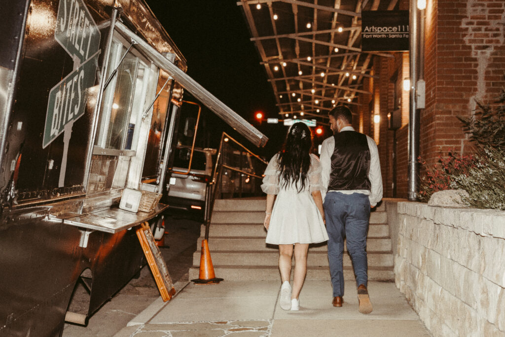 A wedding couple walking hand in hand past food trucks at their Fort Worth wedding reception at Artspace111, blending casual charm with modern celebration vibes.