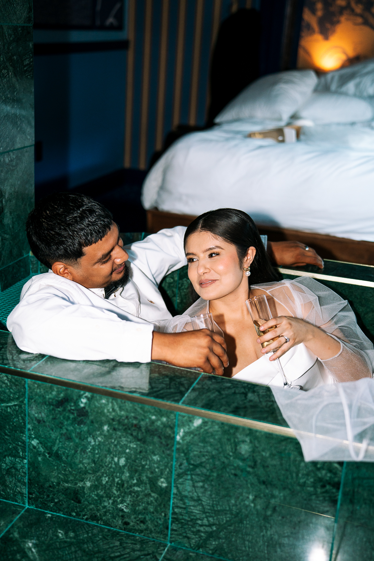 Waco wedding photographer captures newlywed couple at Hotel Herringbone relaxing in a green marble tub, sharing champagne and intimate moments after their wedding.