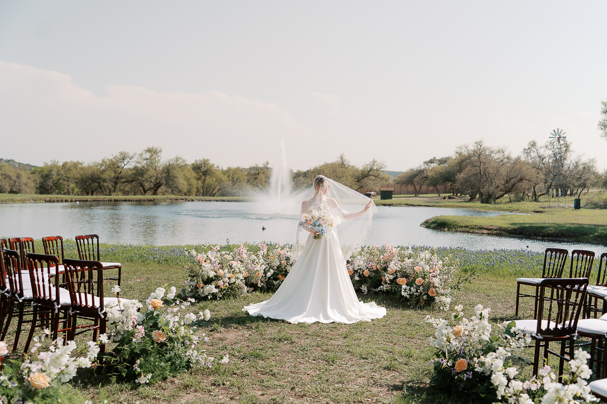 Bride standing in front of a serene lakeside ceremony setup at Ma Maison in Austin, Texas. She holds a pastel bouquet and lifts her veil, surrounded by lush spring florals and wooden chairs, with a fountain and oak trees in the background.