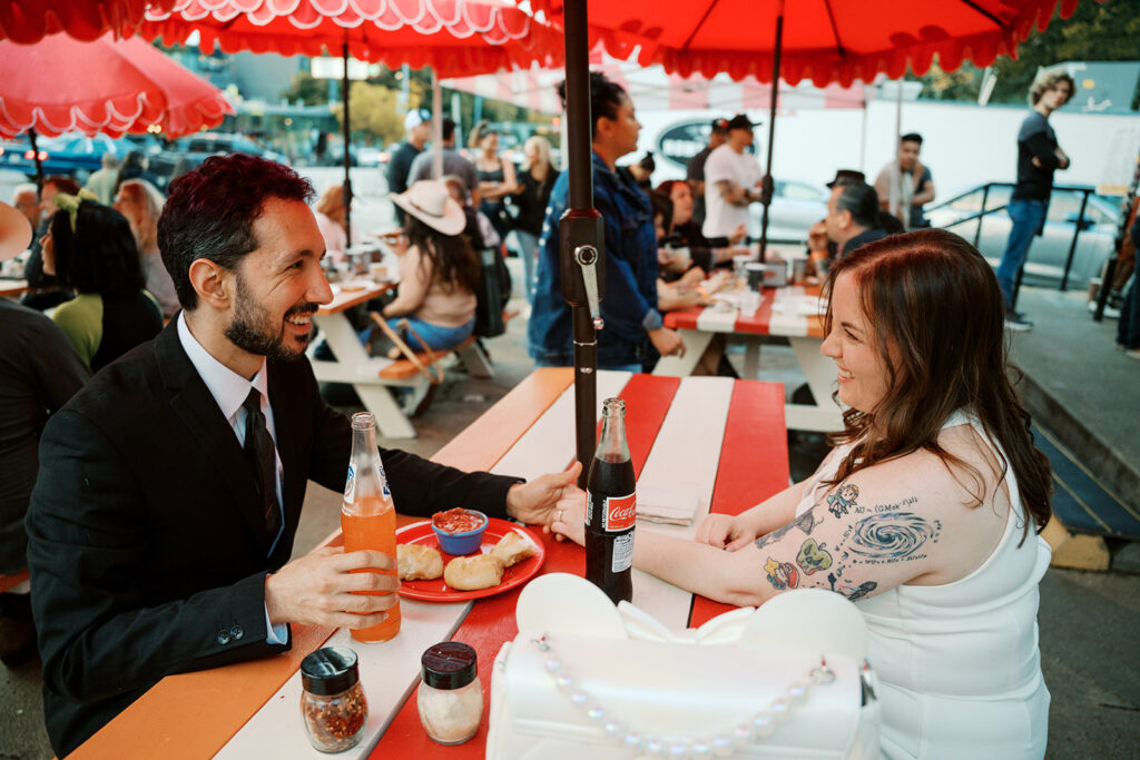 A newlywed couple enjoying bottled sodas and pizza bites under red umbrellas at Home Slice Pizza in Austin, TX. The groom is in a black suit, and the bride, in a white dress with colorful arm tattoos, smiles while they hold hands across a red and white picnic table.