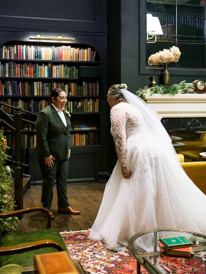 Waco wedding photographer captures emotional first look at Hotel 1928 with an LGBTQ+ couple — one in a lace bridal gown and veil, the other in a dark green suit, surrounded by bookshelves and holiday décor.