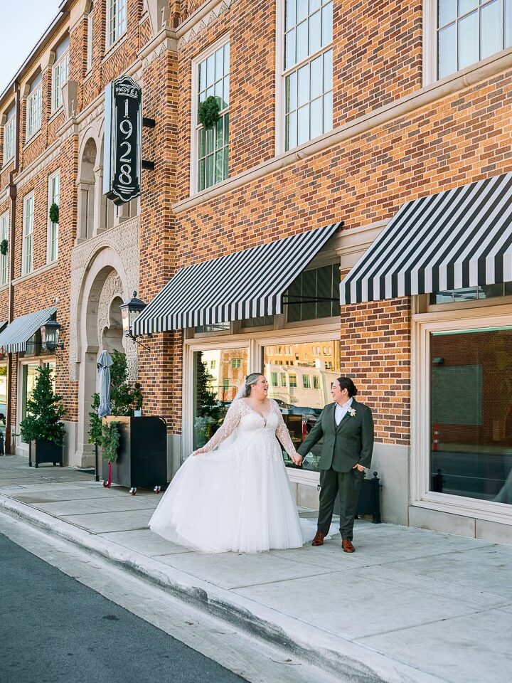 Waco wedding photographer captures LGBTQ+ couple holding hands outside Hotel 1928, with the bride in a lace gown and the partner in a green suit standing in front of the brick building with striped awnings.