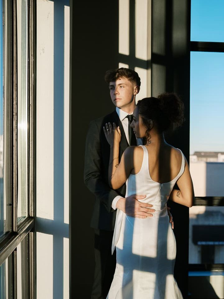 Waco wedding photographer captures a romantic moment at Hotel Herringbone — bride in a satin gown and groom in a black suit embracing by tall windows, with golden hour light casting dramatic shadows.