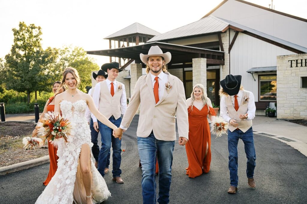 Waco wedding photographer captures a joyful Woodway Arboretum wedding — bride in a lace gown with a slit and boho bouquet holding hands with groom in cowboy hat, blazer, and jeans, surrounded by wedding party in rust orange dresses and western attire.
