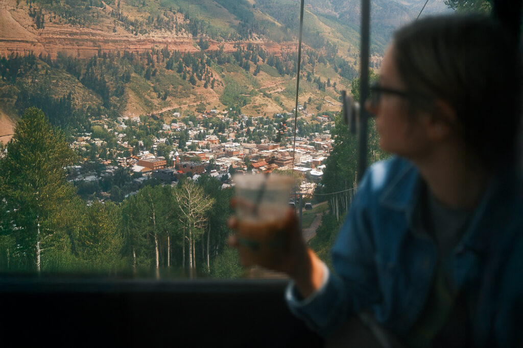 View from a gondola ride overlooking Telluride, Colorado, with a person holding an iced coffee in the foreground and the San Juan Mountains rising in the distance.